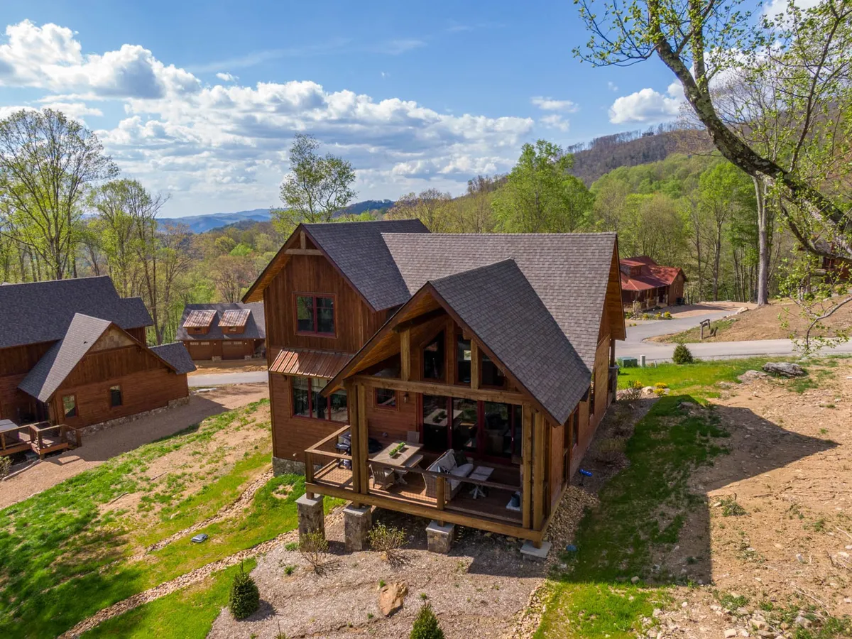 The exterior of a wooden house featuring a large porch with outdoor seating, surrounded by vibrant greenery. The porch is arranged for gatherings and relaxation, providing a seamless indoor-outdoor living experience and a peaceful view of the landscape.