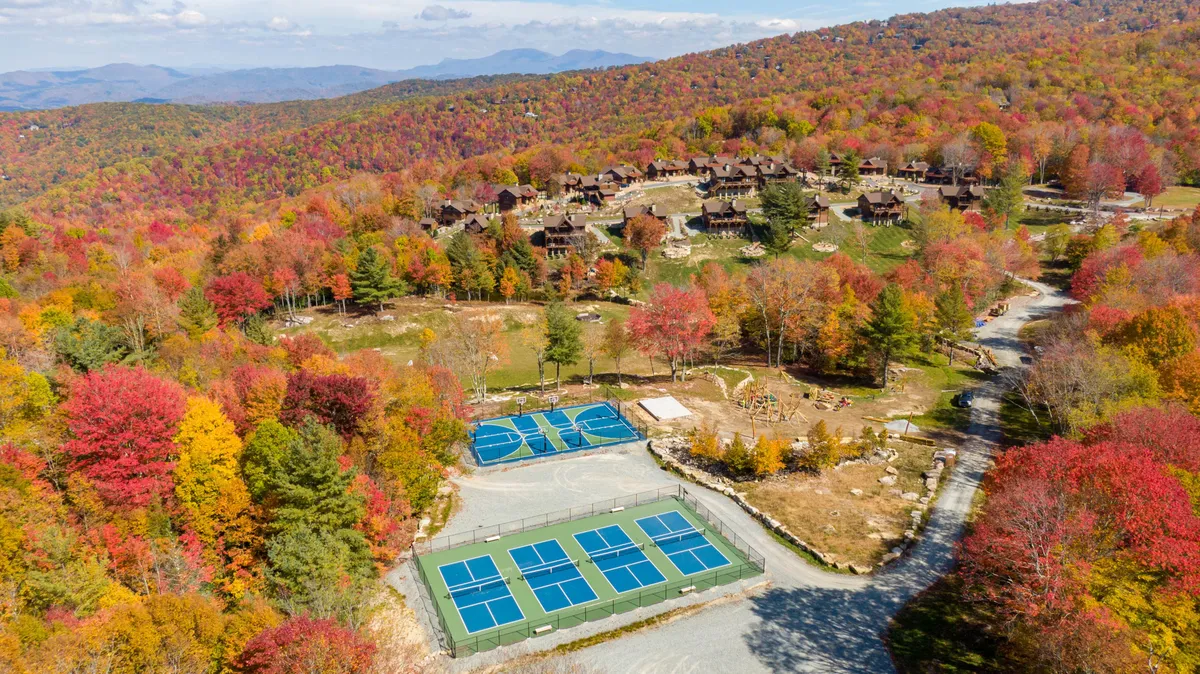 A vibrant autumn scene with colorful foliage framing tennis courts and a mountain view. Cabins nestle in the background, and the area is alive with activity, hinting at sports tournaments, nature walks, and community gatherings typical of family-friendly resorts.