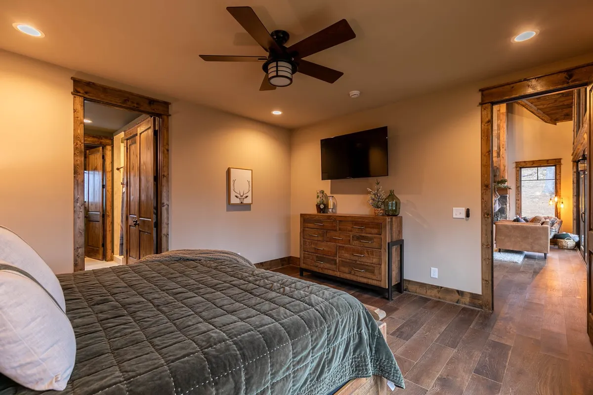 This interior shot depicts a bedroom with a quilted bed, wooden dresser, mounted TV, and a doorway leading to the living area. It offers a glimpse of connectivity within the home and illustrates a transition space from communal activities to private relaxation.