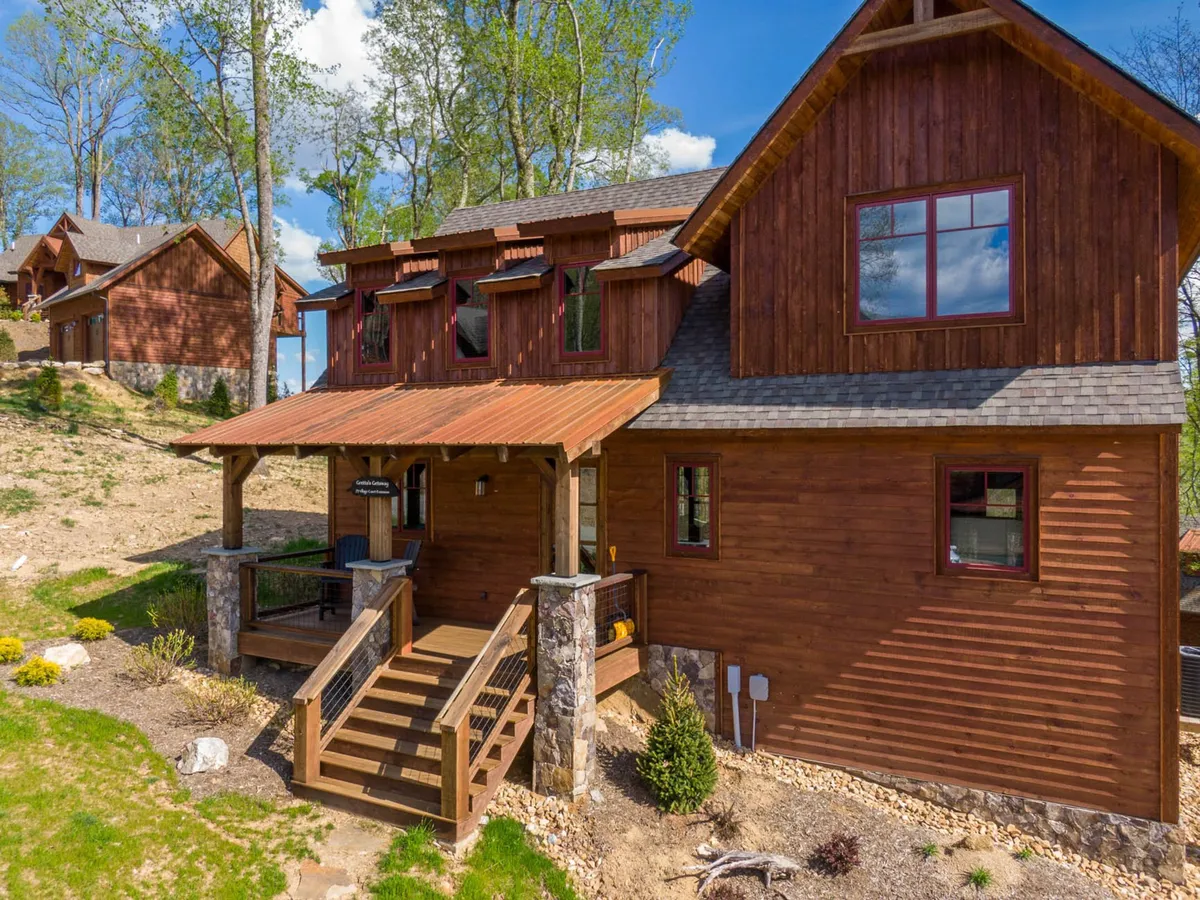 A welcoming exterior view of a wooden mountain home with a spacious porch and stone accents. The porch, reached by stairs, overlooks a serene natural landscape and is surrounded by large windows that invite in natural light. The inviting facade and setting evoke a sense of connection to the outdoors and peacefulness.