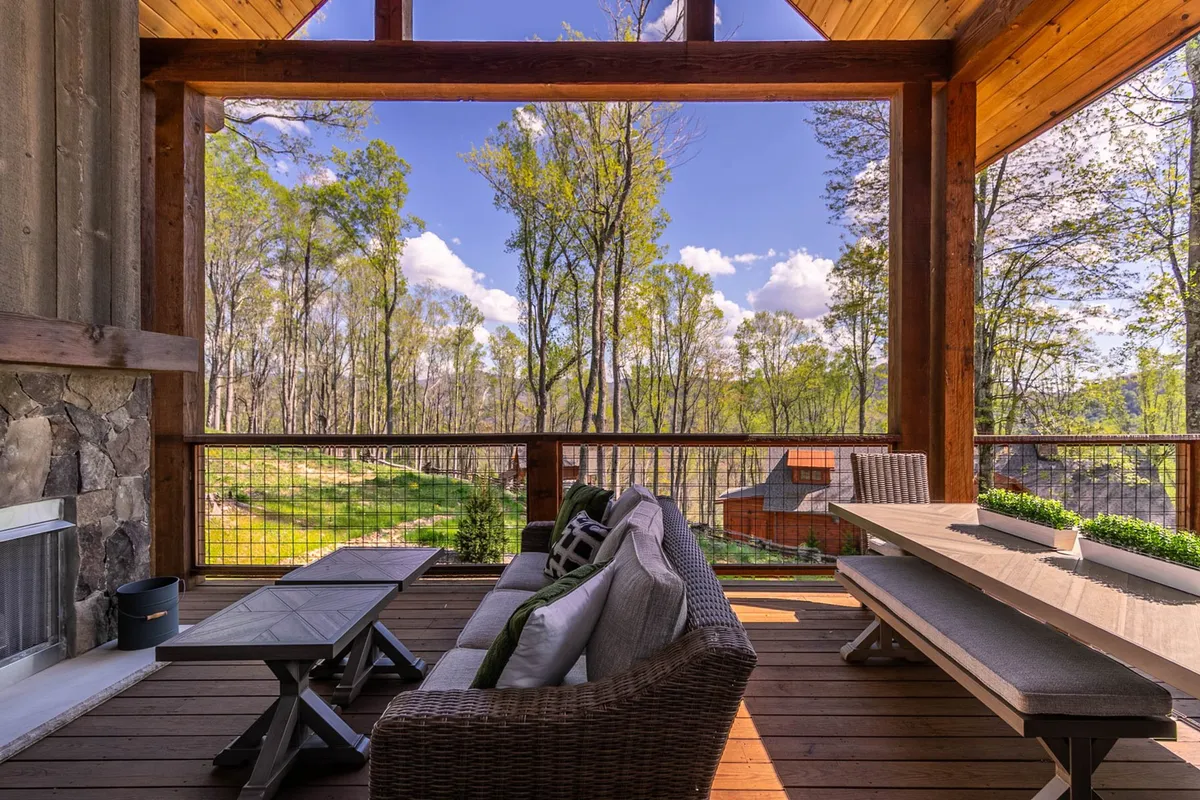 A spacious outdoor deck with a comfortable seating area, dining table for alfresco meals, and a fireplace as a focal point. The deck overlooks serene natural scenery, including trees and distant mountain views, emphasizing a tranquil and private mountain retreat.