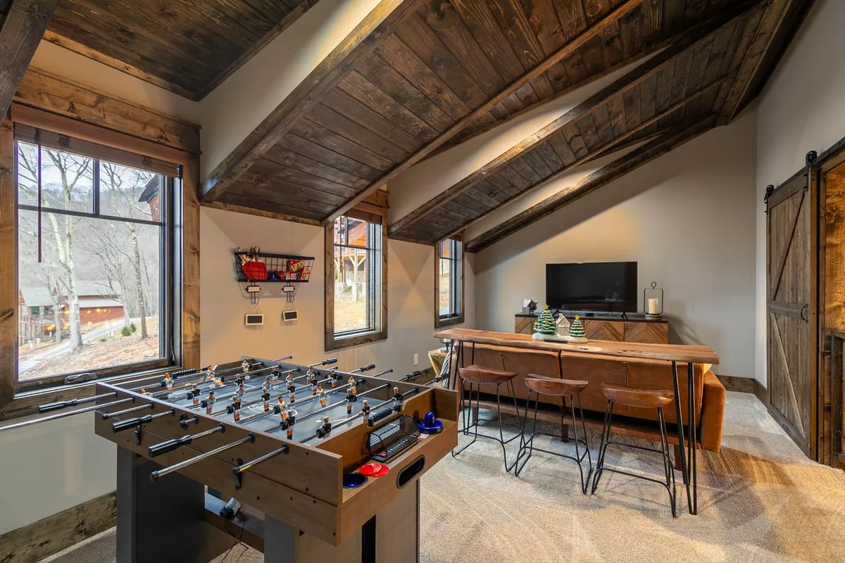 This image features a lively interior space with a foosball table in the foreground, a bar counter with stools, a wall-mounted TV, and large windows flooding the room with natural light. The setup exudes a fun, social atmosphere, perfect for group activities and entertainment.
