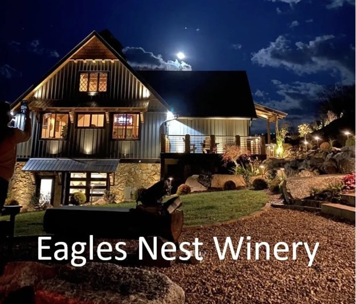 The illuminated facade of Eagles Nest Winery glows warmly against the evening sky, with a landscaped garden in the foreground and the moon overhead. The scene exudes a welcoming atmosphere, inviting guests to enjoy wine tasting and events under the stars.