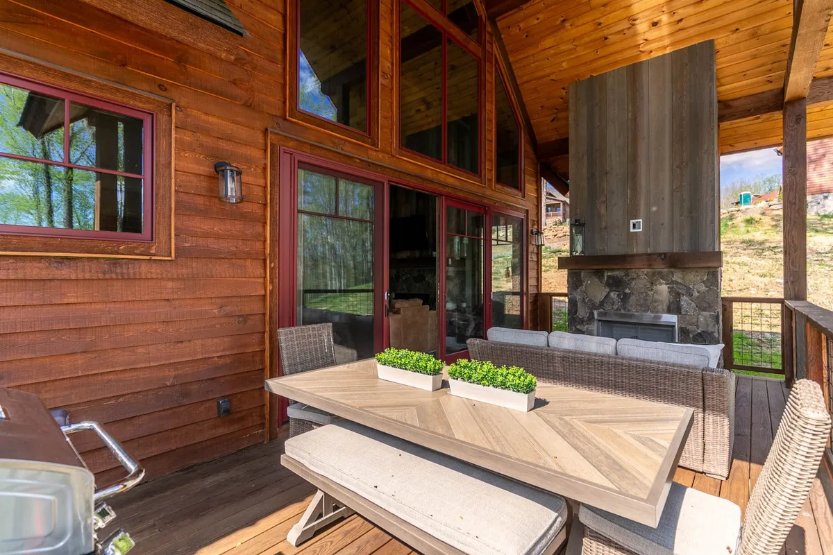 A covered outdoor living space featuring a substantial stone fireplace, a wooden dining table with comfortable seating, and large glass windows looking into the home. The scene is bathed in warm, ambient lighting, suggesting an inviting setting perfect for both day and evening use.