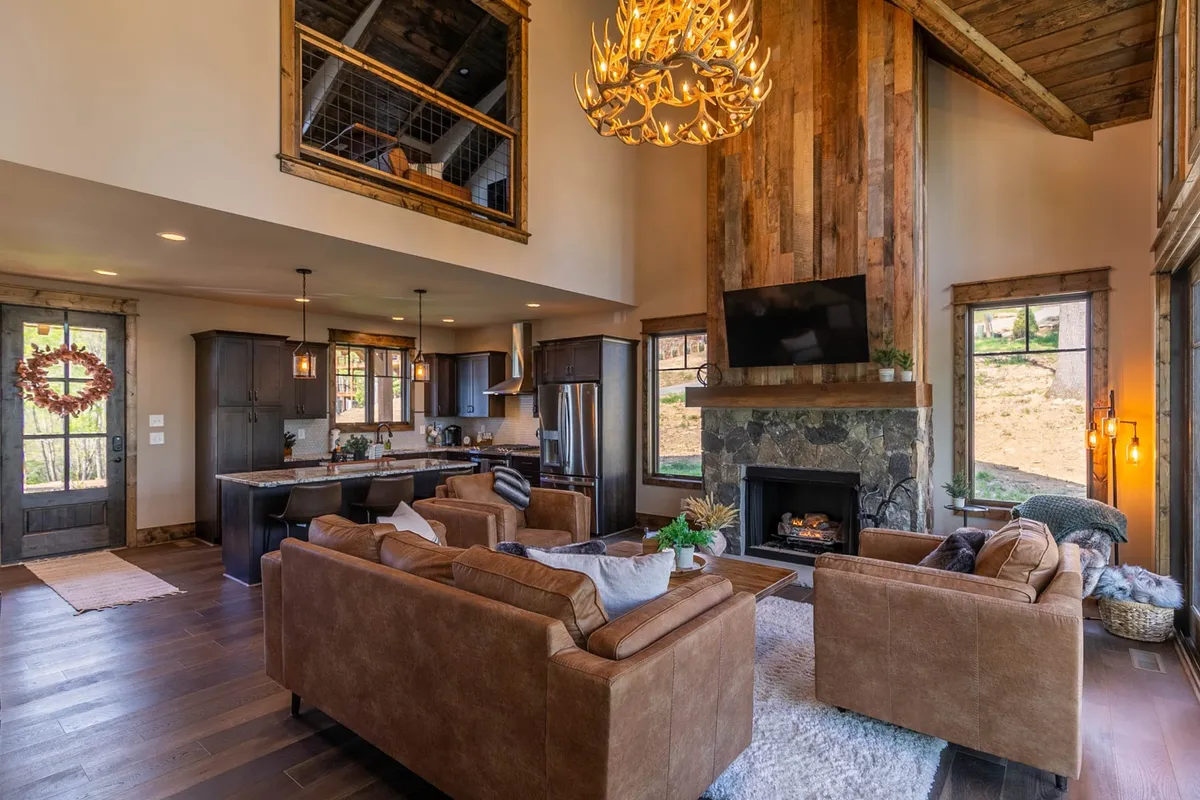This image features a spacious living area with comfortable leather sofas, a large stone fireplace, an antler chandelier, and expansive windows offering striking mountain views. The open layout flows naturally into the kitchen area, providing a sense of togetherness and warmth.