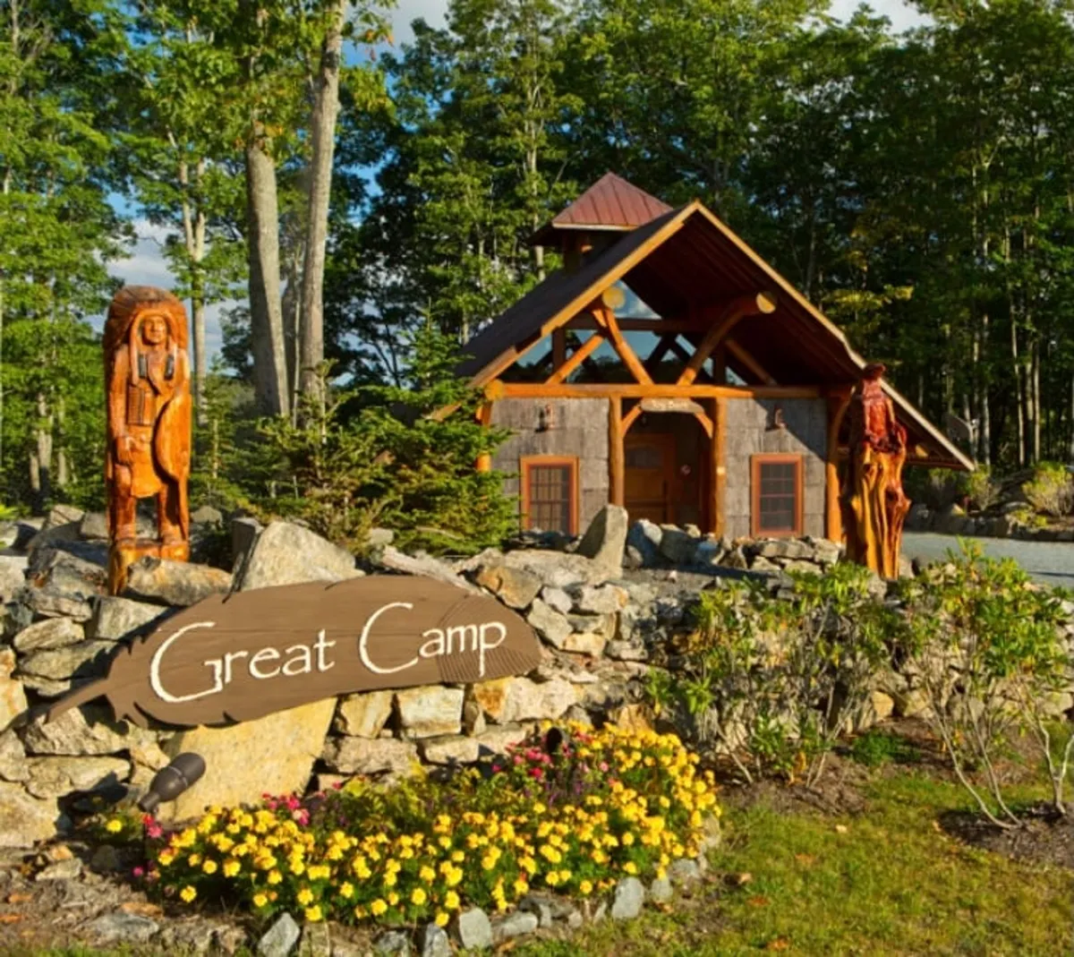 A lively outdoor scene featuring a rustic log cabin structure, the 'Great Camp' sign, a striking wooden sculpture, and vibrant flower beds. The space suggests communal outdoor activities, gathering spots, and artistic touches, set against a natural backdrop.